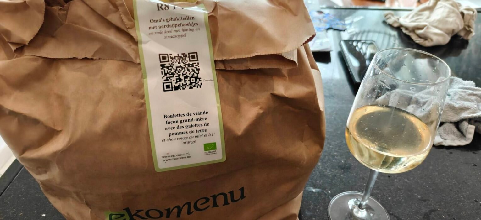 Papieren zak van Ekomenu met het label Oma's gehaktballen met aardappelkoekjes, met een glas witte wijn ernaast